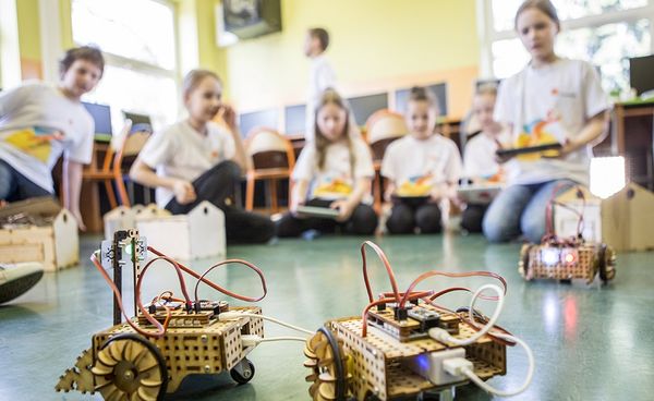 Na pierwszym tle dwa jeżdżące roboty, natomiast na drugim siedzące dzieci z tabletami