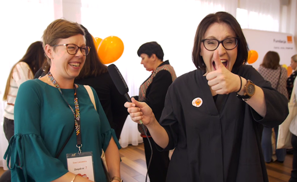 na pierwszym planie dwie kobiety obie uśmiechnięte z czego jedna ma podniesiony kciuk do góry