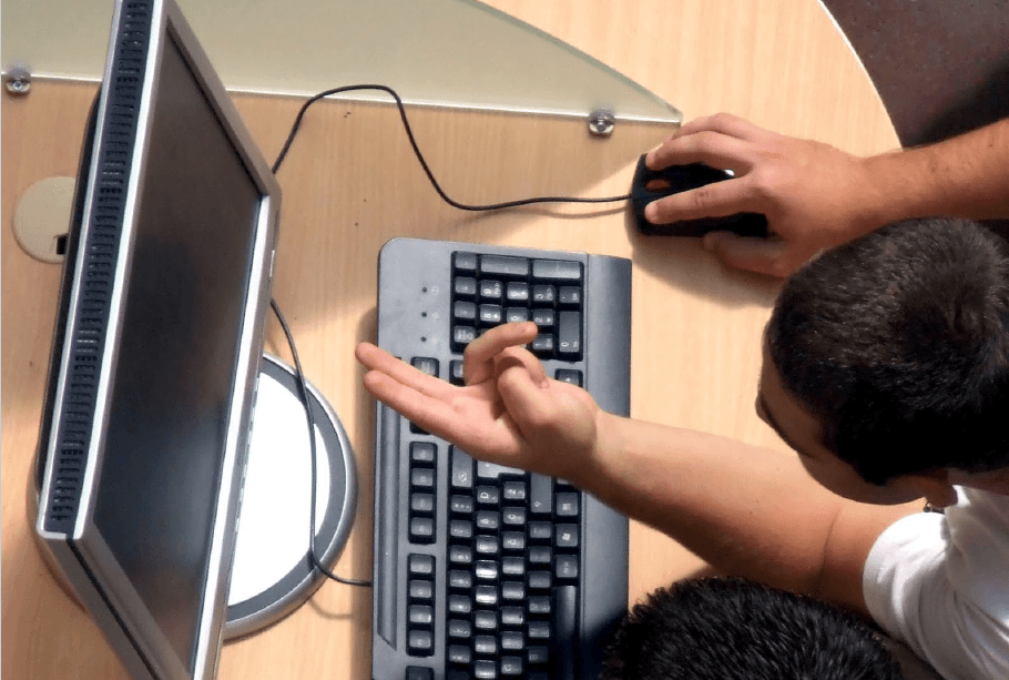 Mężczyzna siedzący przed komputerem w jednym ręku trzyma myszkę od komputra, natomiast drugą wskazuje na ekran monitora
