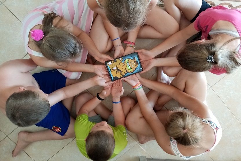 Grupa dzieci siedząca w kółku z połączonymi rękami przed sobą trzymając tableta