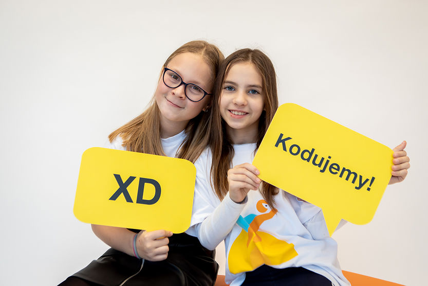Dwie dziewczynk siedzące z tabliczkami z napisami "XD" oraz "Kodujemy!"