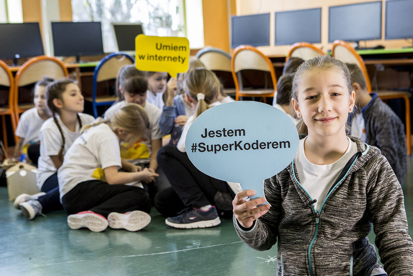Zdjęcie dziewczynki trzymającej tabliczke z napisem "Jestem #SuperKoderem" na tle grupki siedzących dzieci